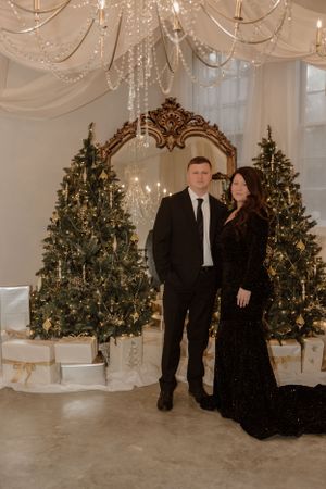 Formal couple portrait in front of Christmas trees and ornate mirror with chandelier overhead.