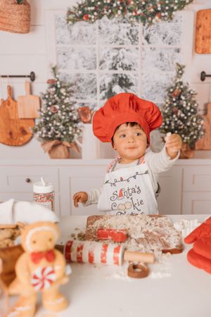 A young baker wearing a red chef hat and apron making Christmas cookies in a festive kitchen.
