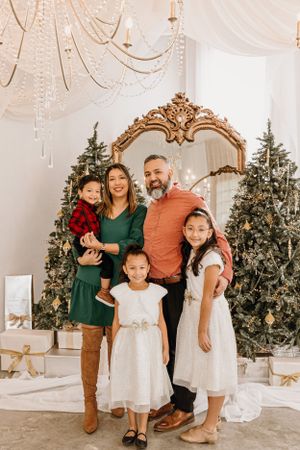 Holiday family portrait with decorated Christmas trees and gold mirror in elegant white room setting.
