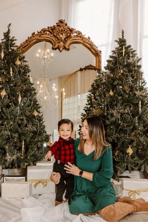 Mother and child share joyful moment by Christmas trees and ornate gold mirror during holiday photoshoot.