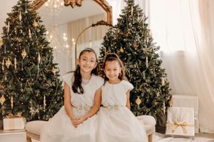 Two children in white dresses pose in front of decorated Christmas trees with a gold ornate mirror and white curtains.