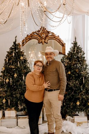 Couple poses in front of decorated Christmas trees with gold mirror and crystal chandeliers.