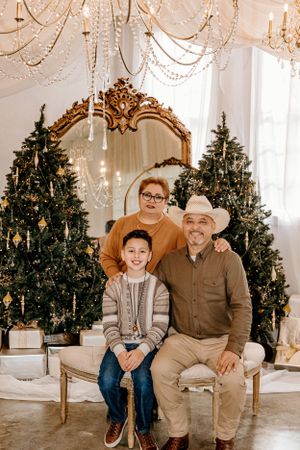 Family of three poses on bench with decorated Christmas trees and ornate mirror in background.