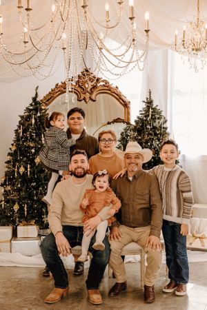 Multi-generational family portrait in front of Christmas trees with ornate gold mirror and chandeliers.