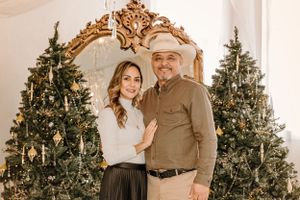 Couple poses together in front of decorated Christmas trees and gold ornate mirror.