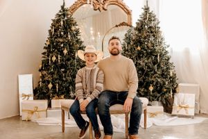 Parent and child sitting on bench in front of Christmas trees for holiday portrait.