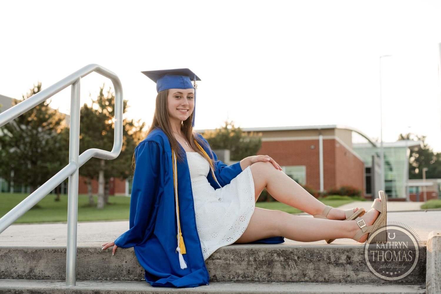Graduate in blue cap and gown sits on concrete steps wearing a white dress.