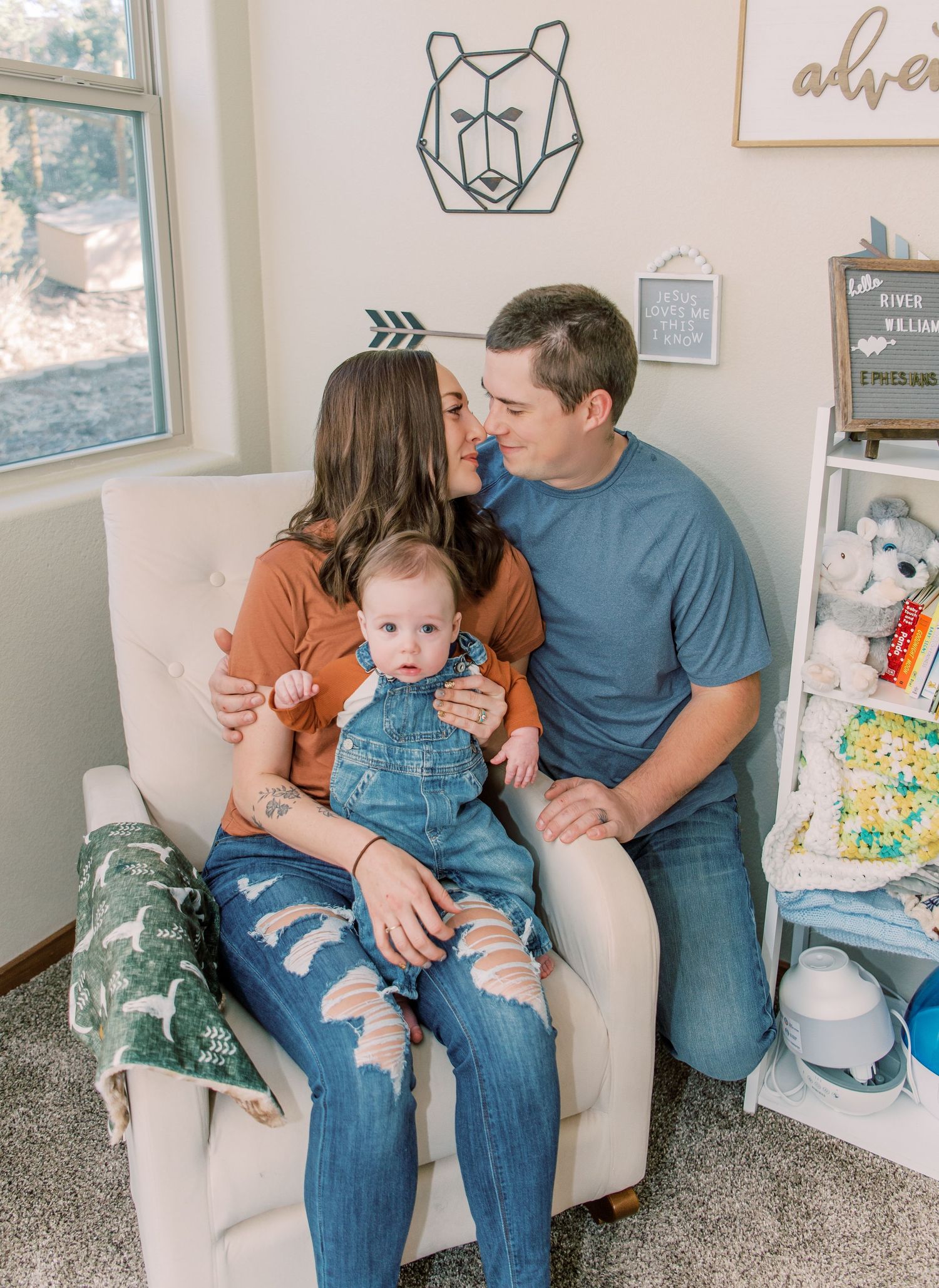 A family cuddles together on a light gray armchair in a nursery room decorated with a geometric bear wall art.