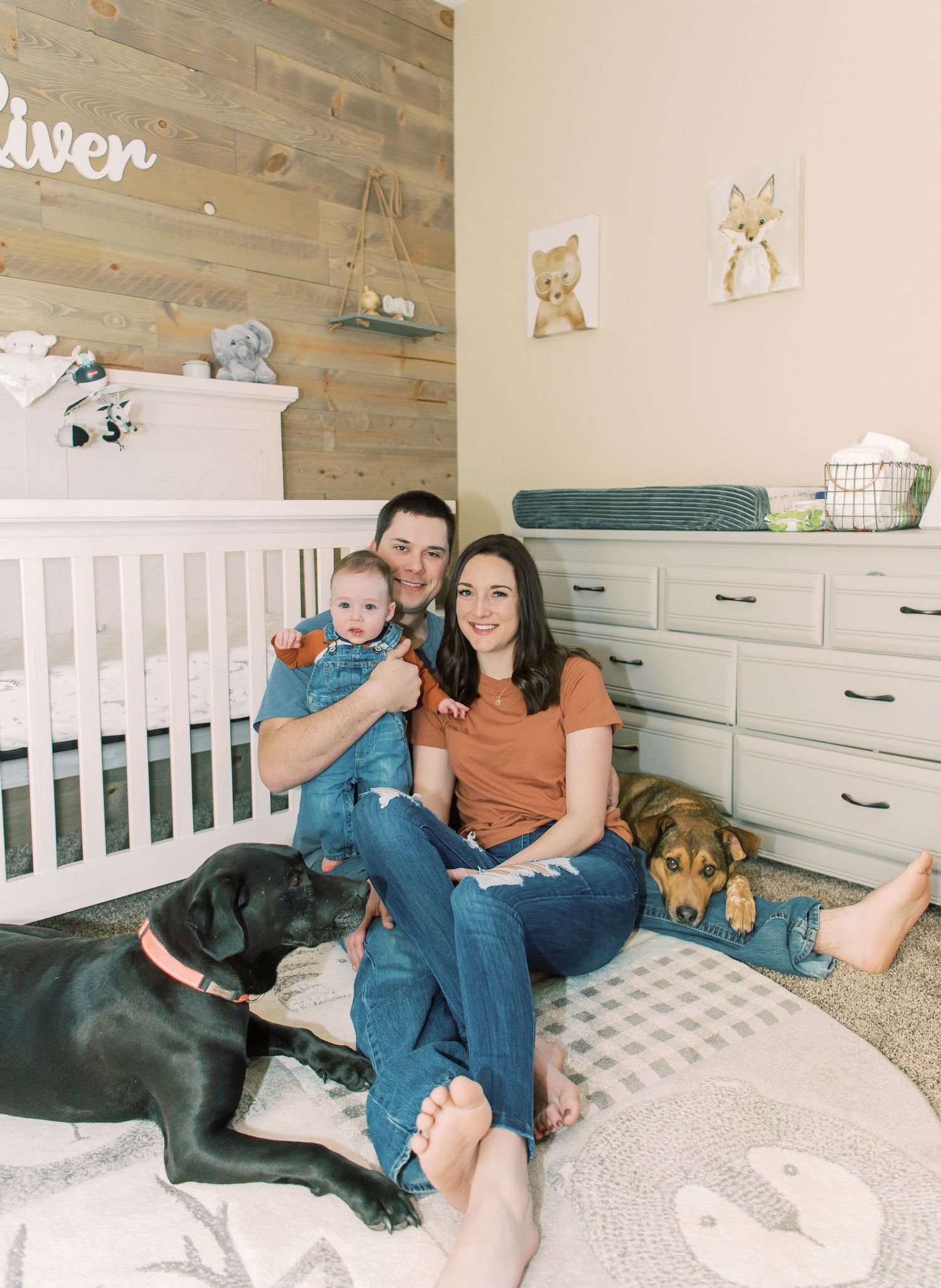 A cozy nursery room with wooden accent wall features a white crib, dresser, and two dogs relaxing on the floor.