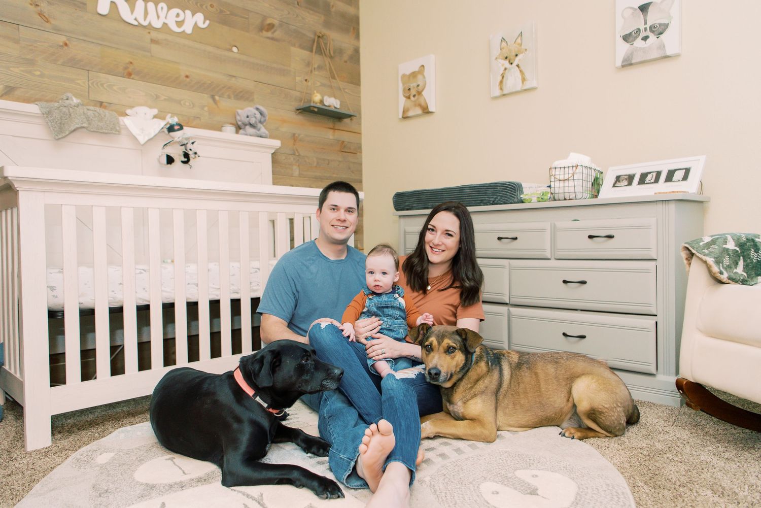 A nursery room with a white crib, gray dresser, decorative wall art, and two dogs laying next to a family on the floor.