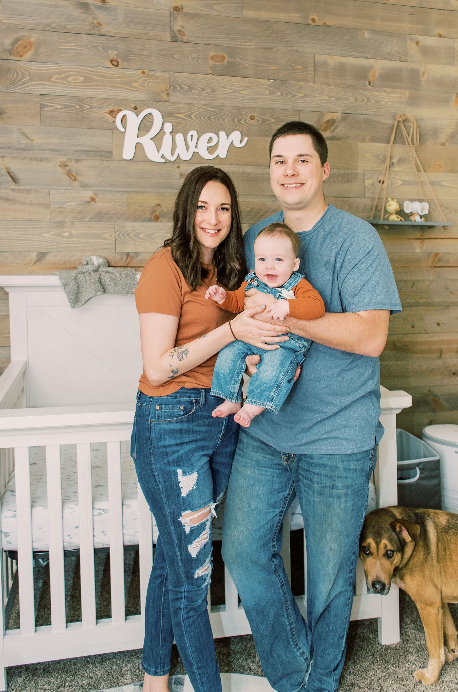 A family poses in a nursery room with wooden walls, a white crib, and a dog sitting nearby.
