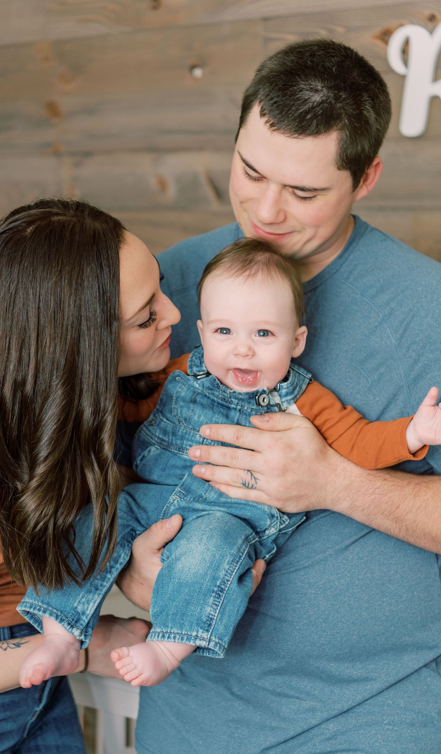 Happy family sharing tender moment with smiling baby wearing denim outfit against wooden wall background.