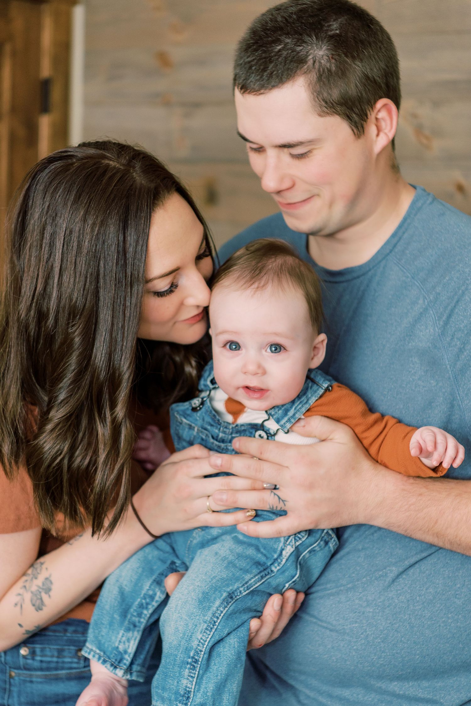 A young family shares an intimate moment together, with a baby wearing denim overalls and an orange shirt.