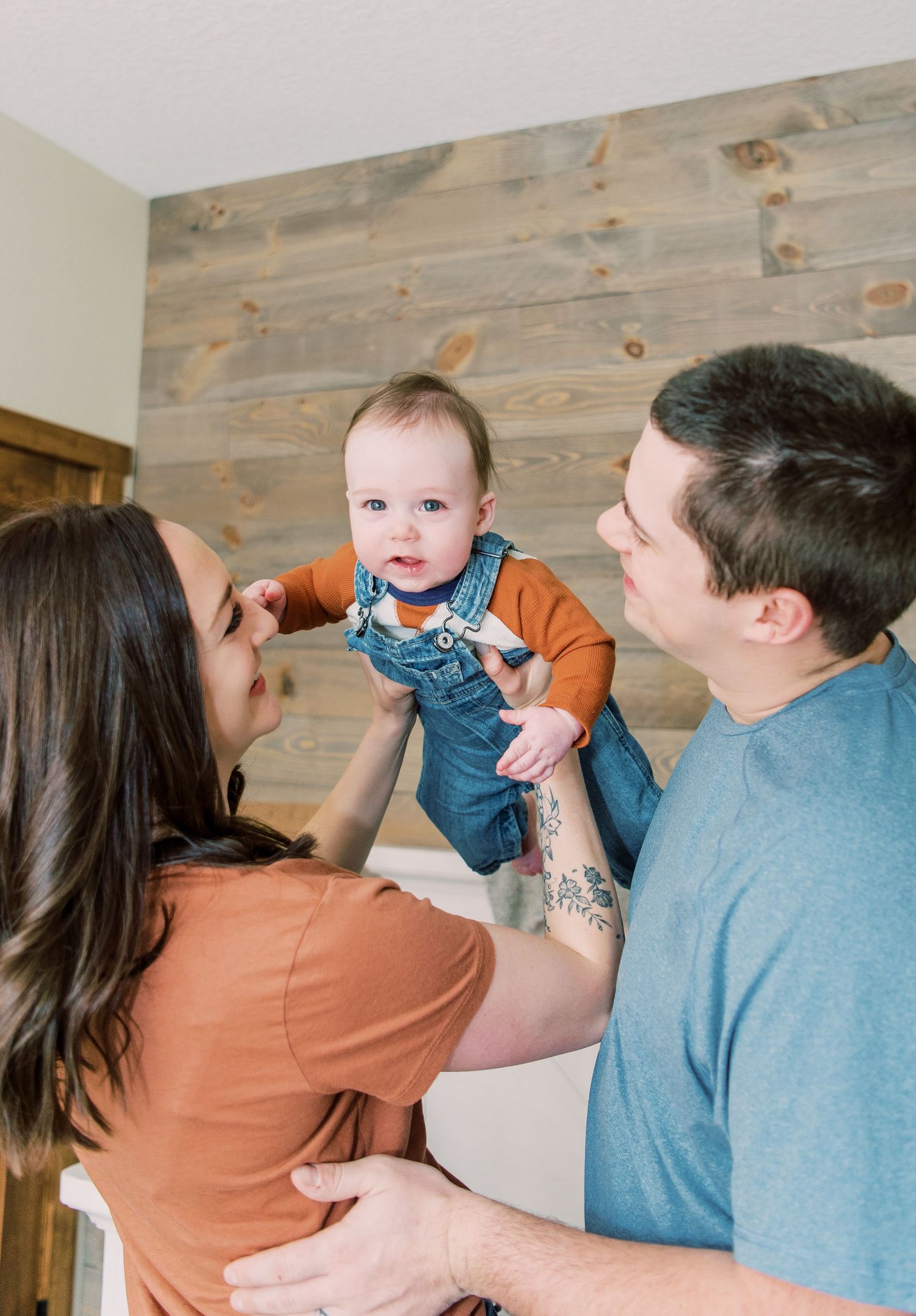 A baby wearing blue overalls and orange shirt is lifted up by two adults against a wooden plank wall.