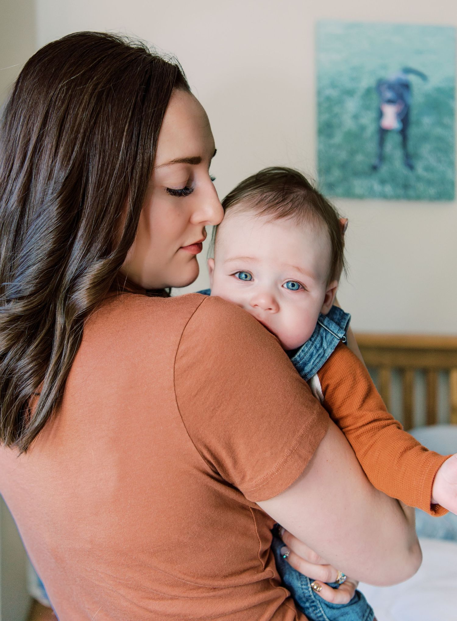 Mother in terracotta shirt holds and lovingly looks at baby wearing blue and orange while at home.