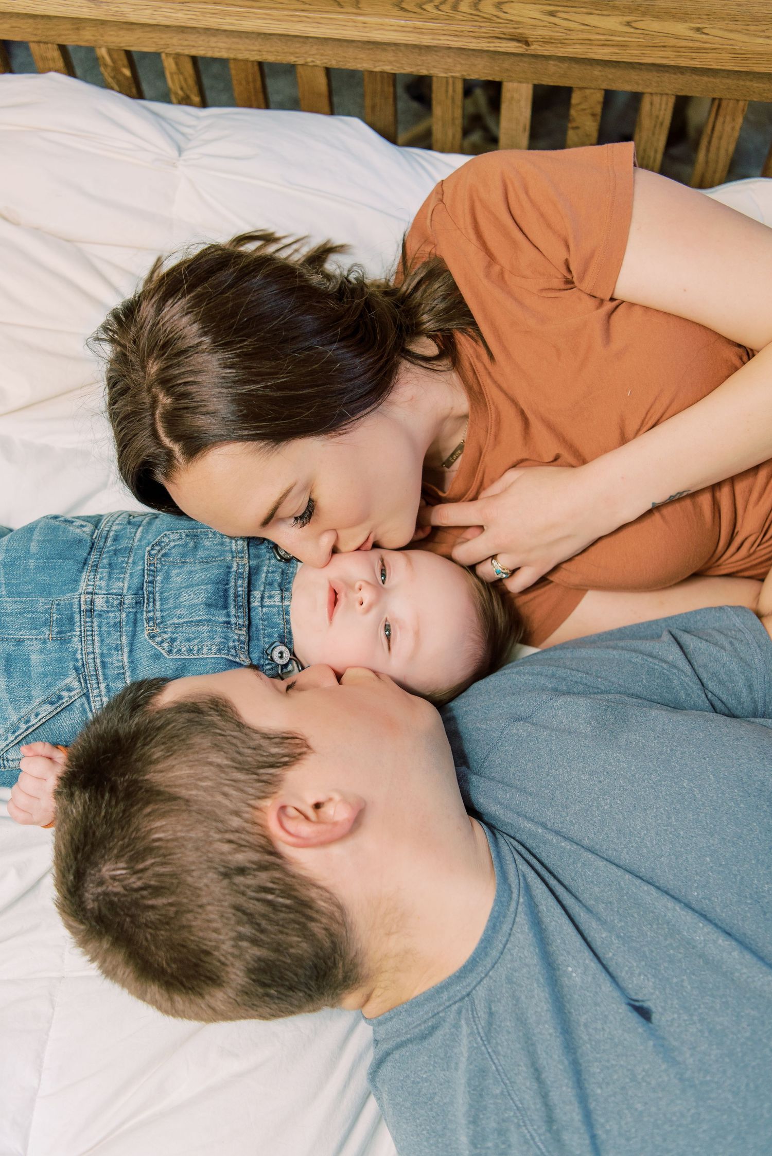Family cuddles together on white bedding, sharing an intimate moment of affection in natural light.