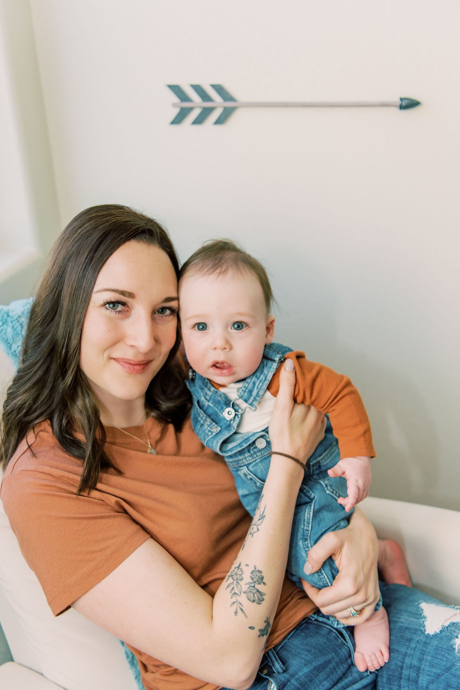 A parent and baby wearing coordinating orange and denim outfits sit together in a chair beneath a decorative arrow wall decal.