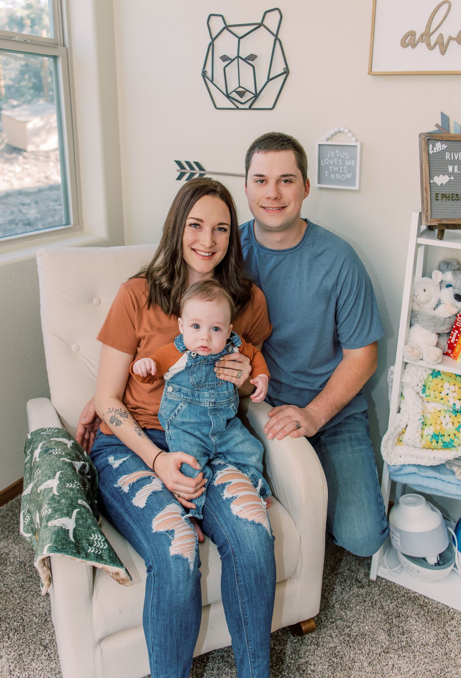Family of three sitting together on beige armchair with geometric bear art on wall in nursery decor.