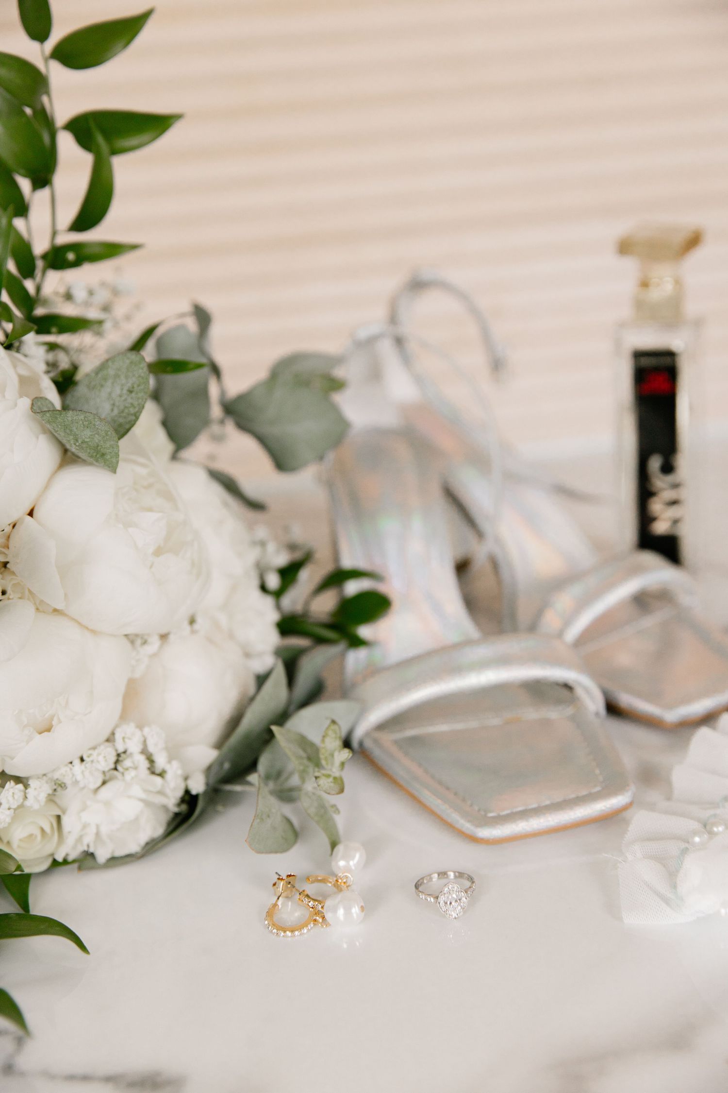 Wedding details with white flowers, silver shoes and jewelry on white surface.