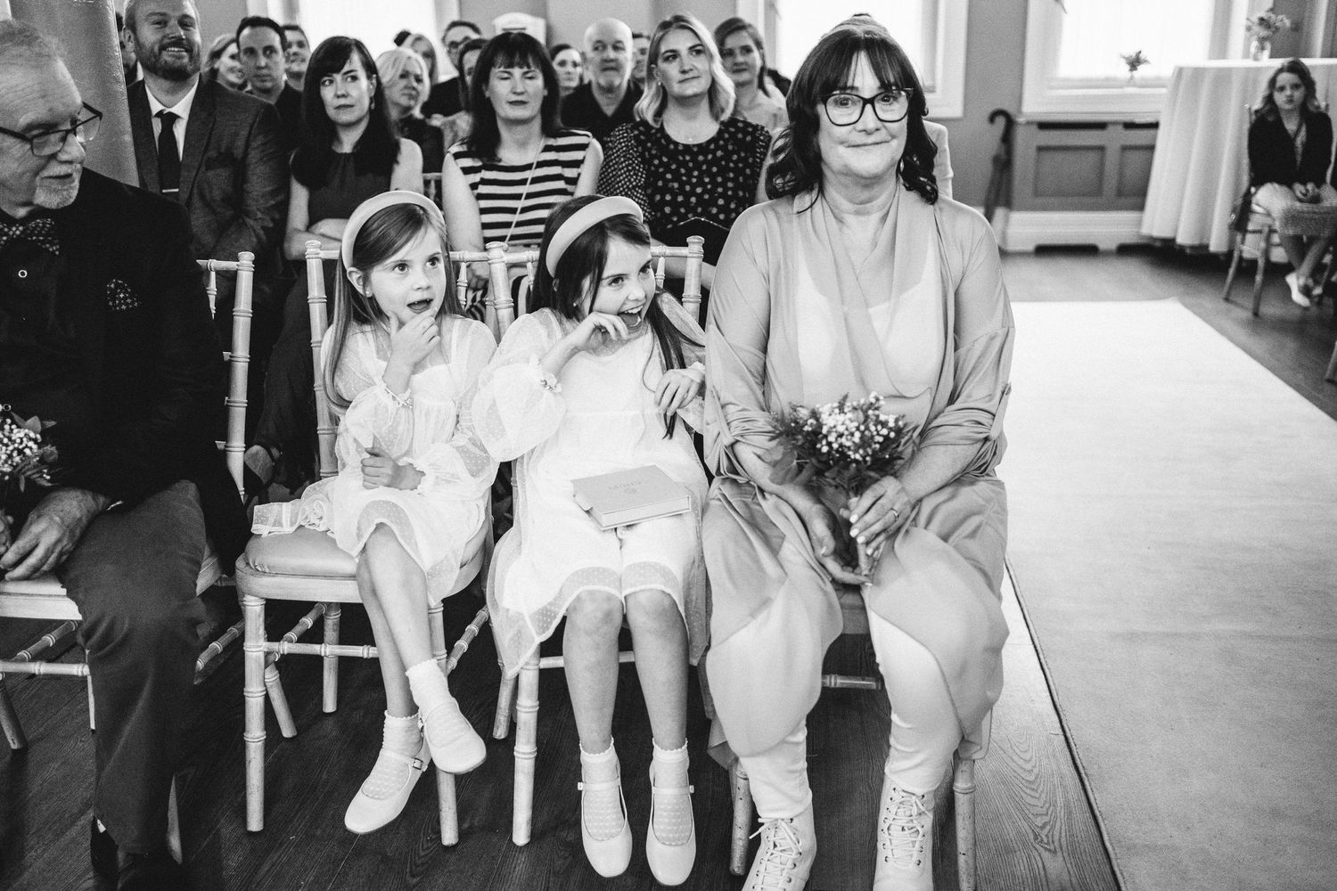 A vintage black and white photograph capturing wedding ceremony guests seated in rows inside a bright venue.