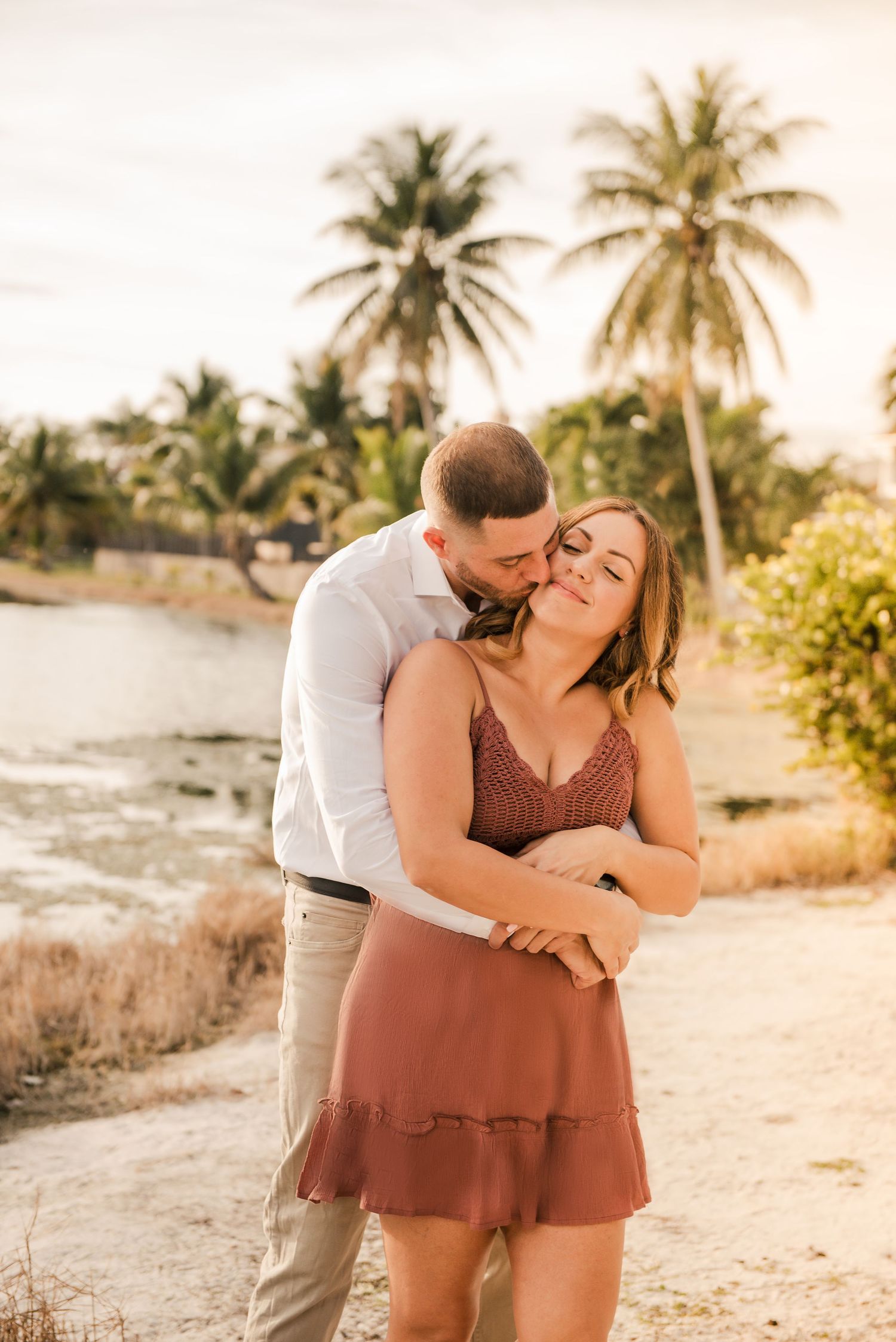 Romantic couple embracing on tropical beach path with palm trees at sunset in warm golden light.