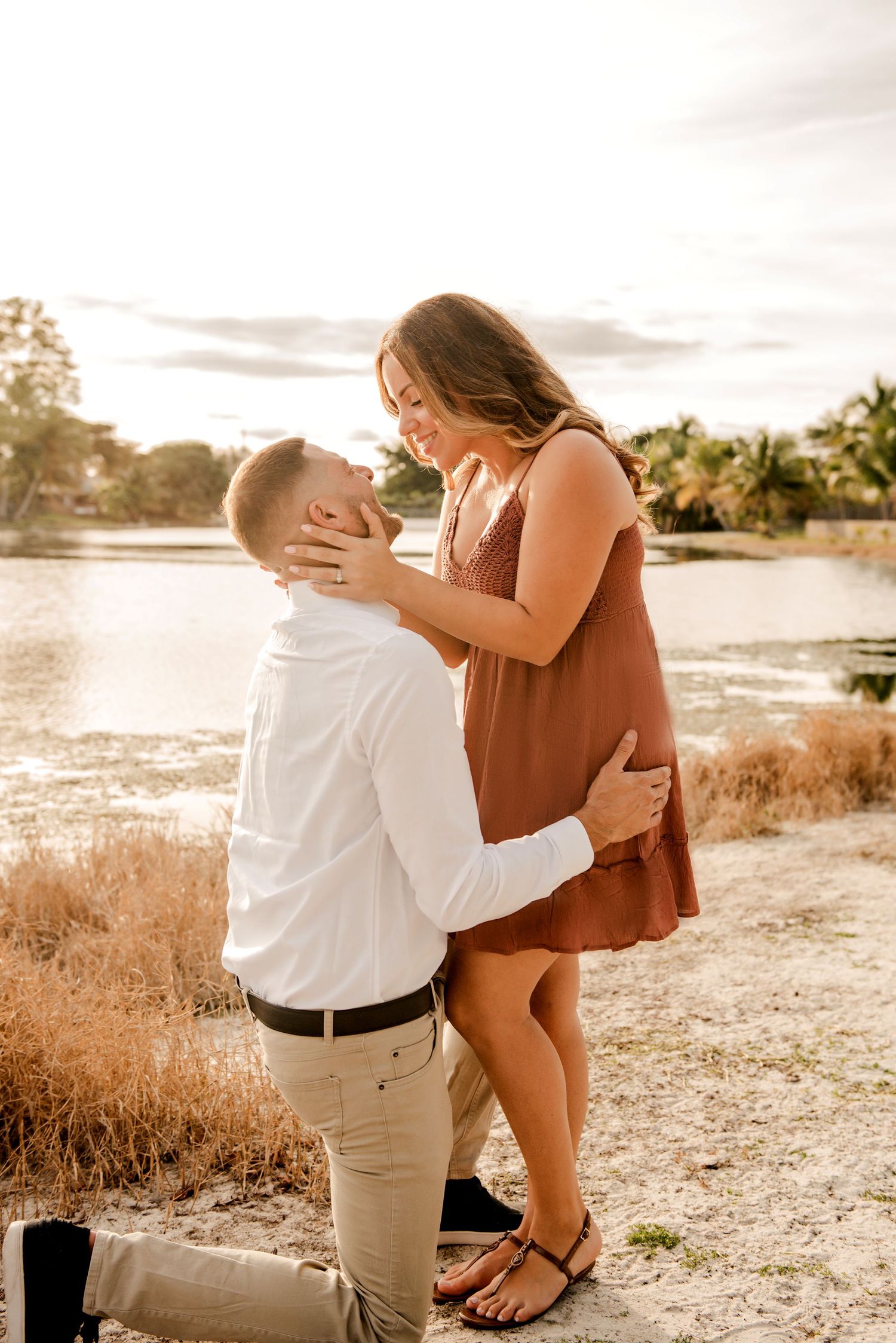 A romantic moment captured during sunset as a couple embraces on a beach with golden light and palm trees in the background.