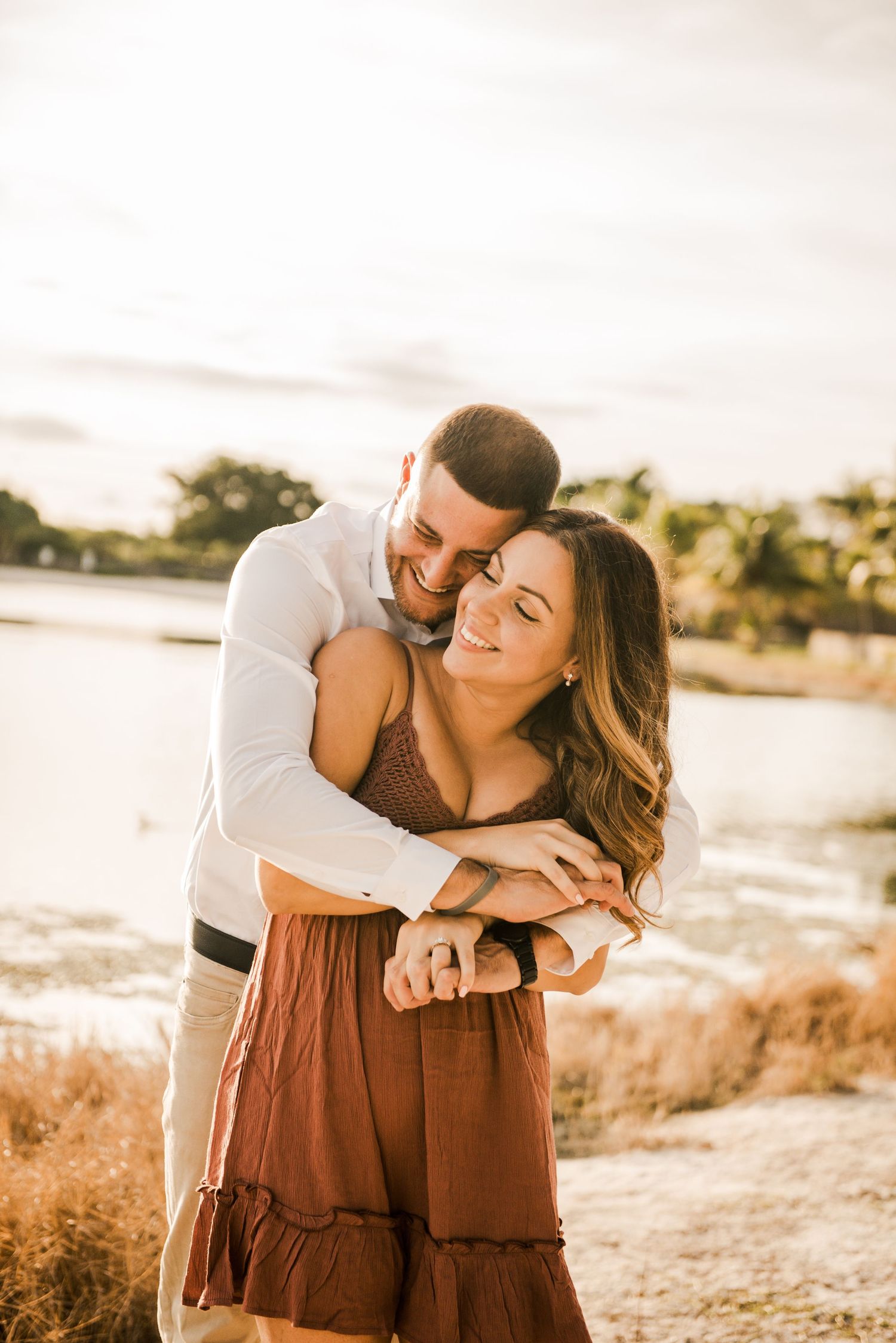 A romantic couple embraces at sunset by a waterfront, with golden light illuminating their intimate moment.
