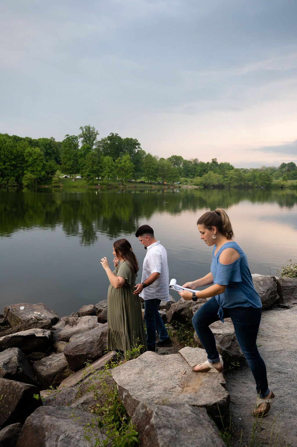 People fishing on rocky shoreline at sunset with calm lake waters and wooded background.