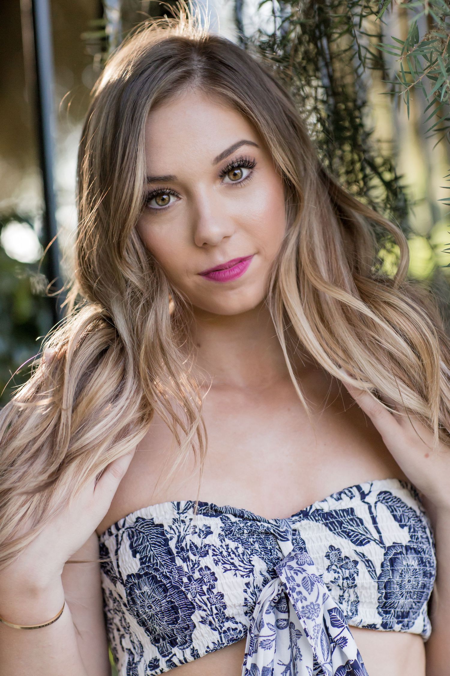 Blonde woman in blue floral strapless top posing outdoors with soft natural background.