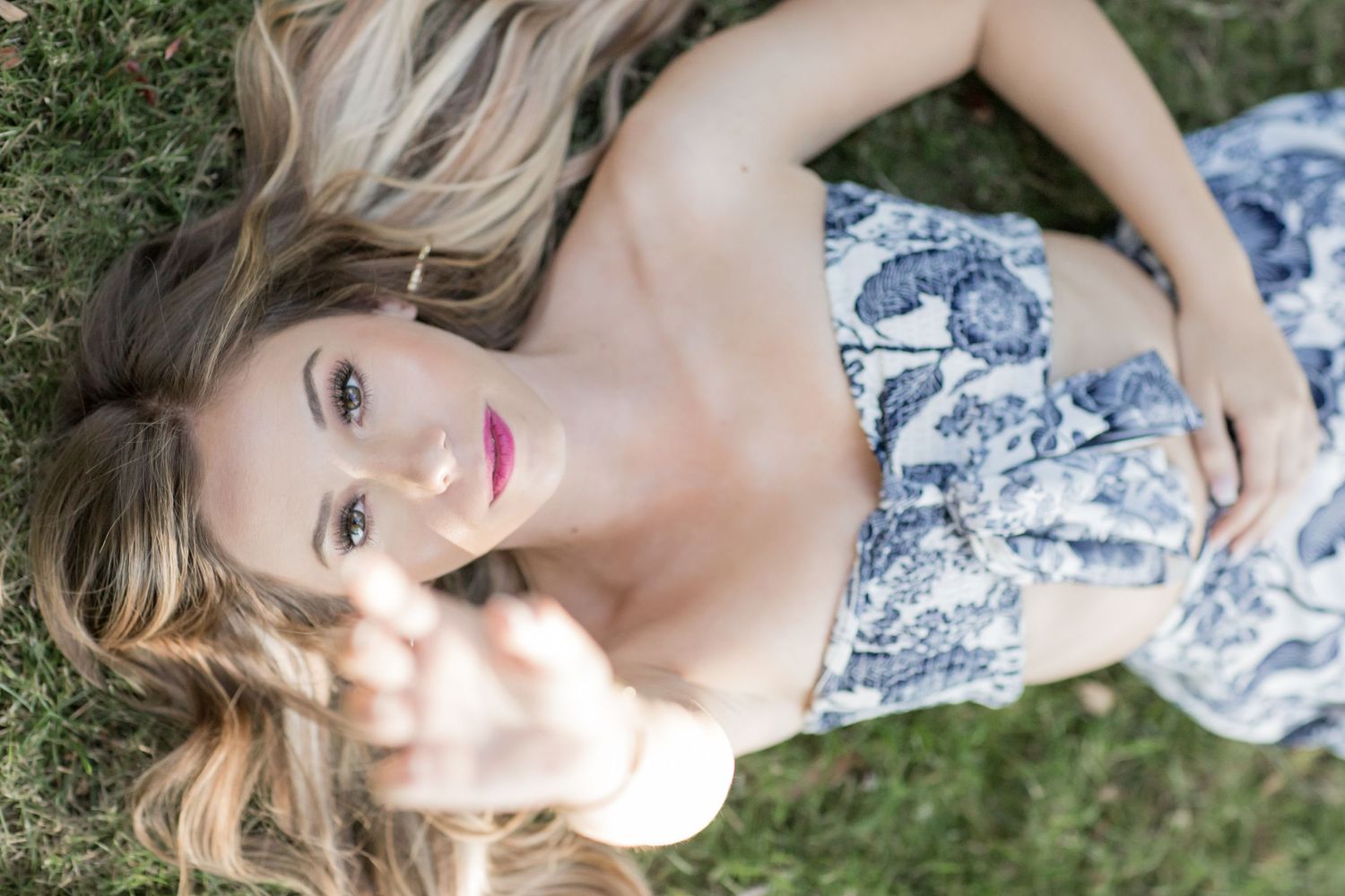 Blonde woman in blue floral dress lying on grass reaching toward camera with pink lips.