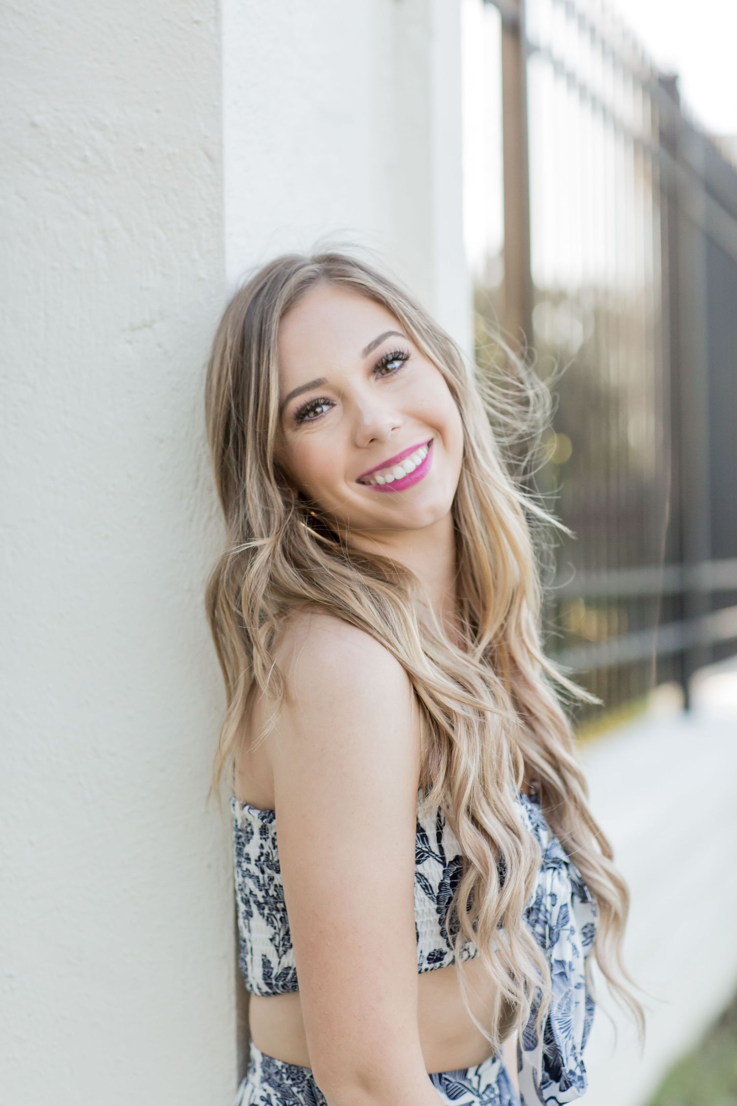 Young woman with long blonde wavy hair smiling against a white wall in a blue floral crop top outfit.