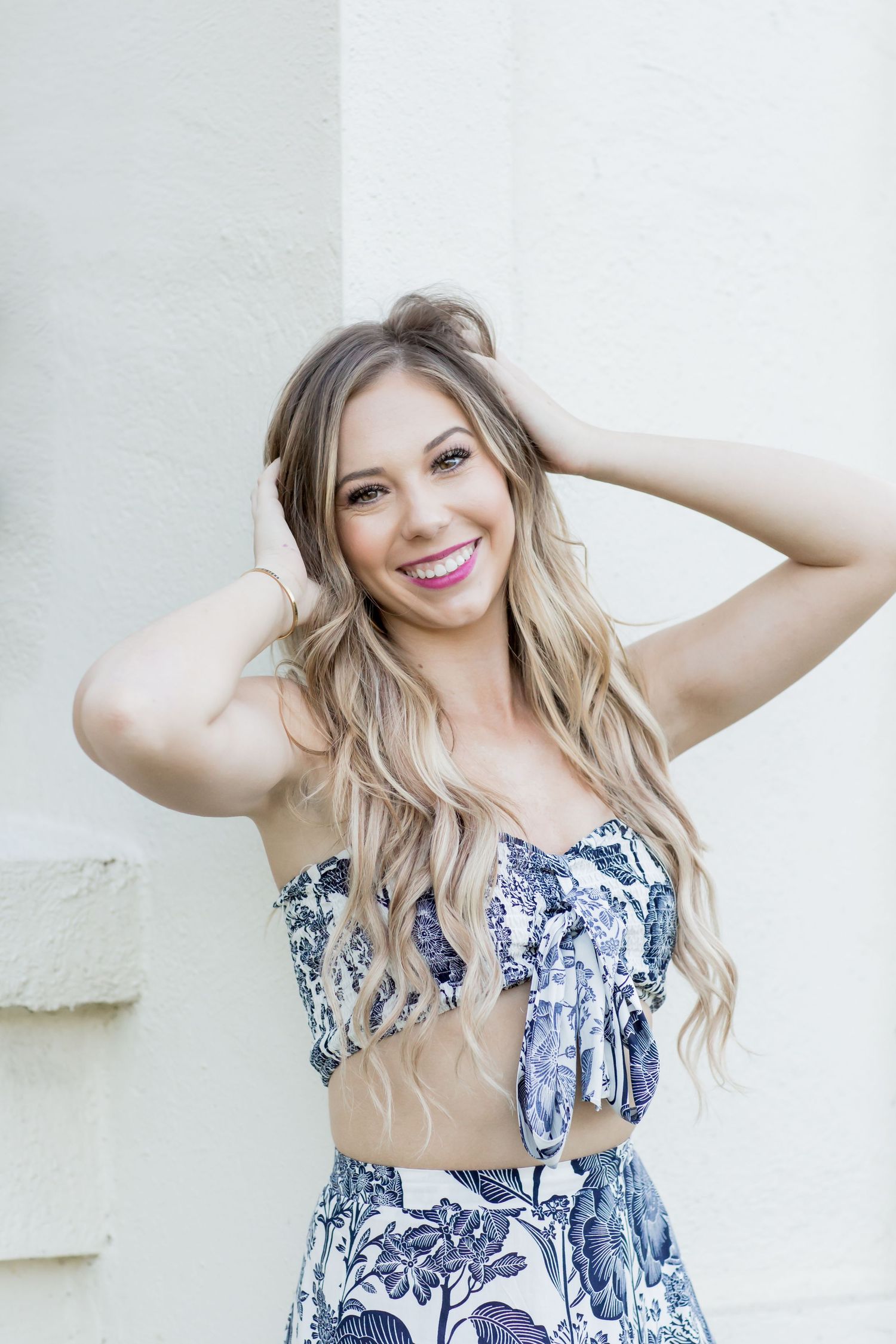 Smiling blonde woman in blue floral two-piece outfit posing with hands in hair against white wall.