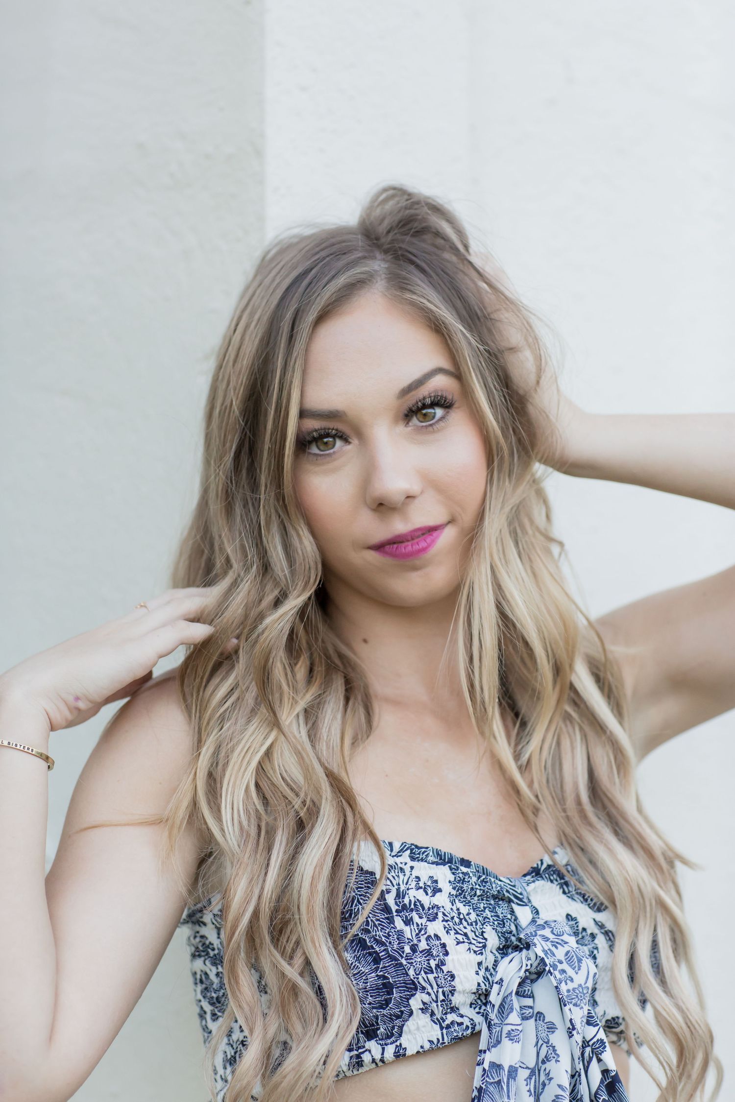 Beautiful blonde woman in blue floral crop top posing with hands in wavy hair against white wall.