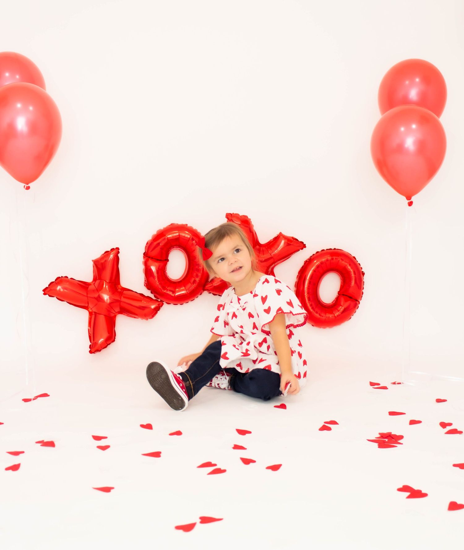 A child sits on white backdrop with red XOXO balloons and heart confetti for Valentine's Day.