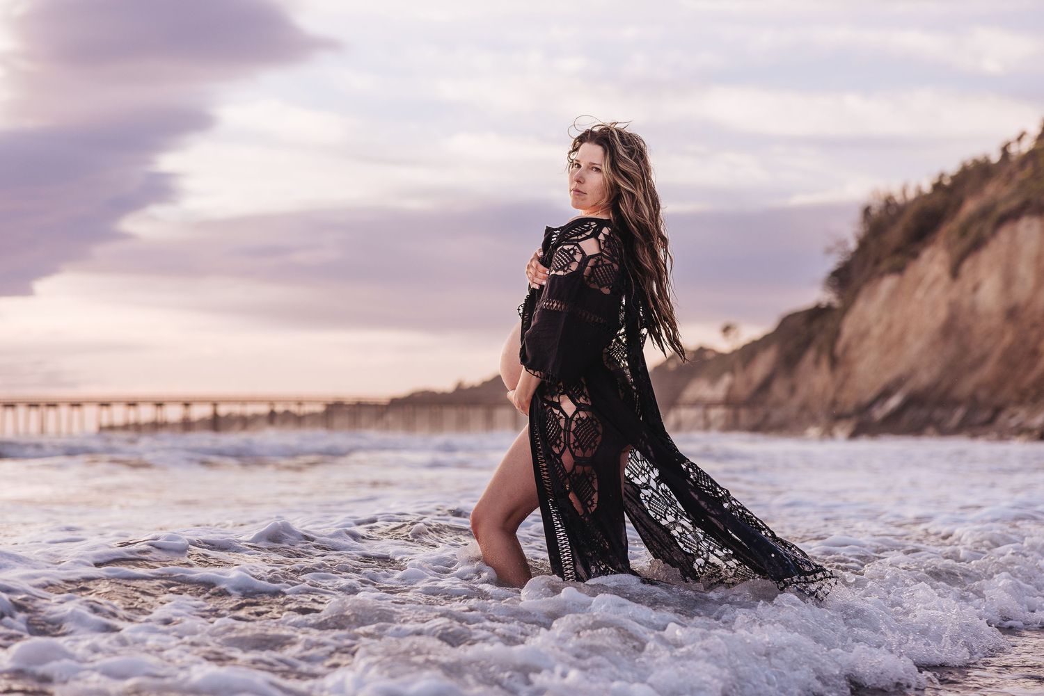Dramatic maternity photo with cliffside and waves at sunset beach.