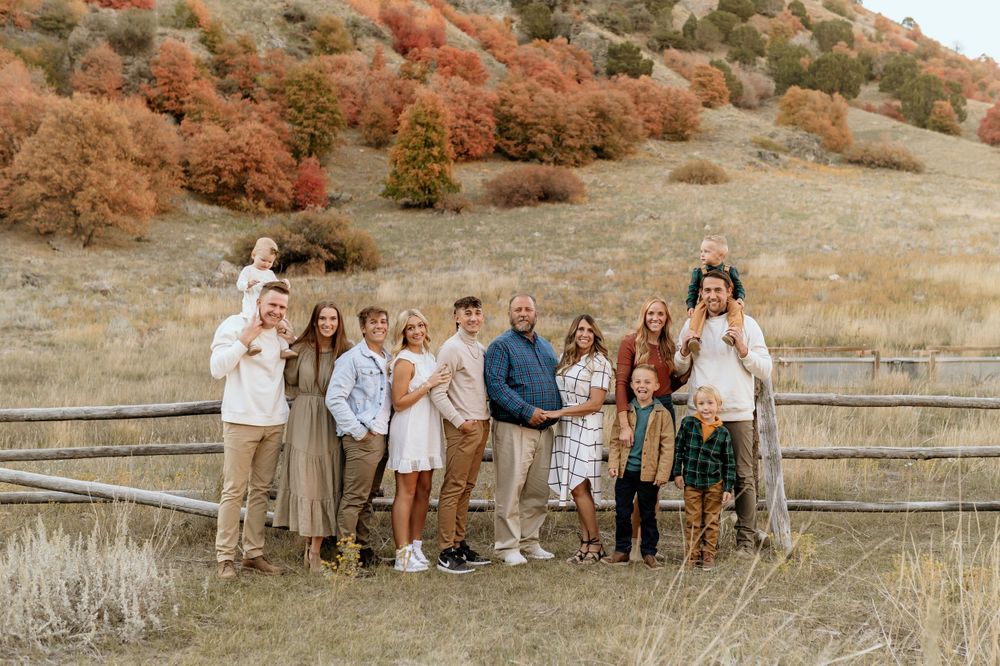 Extended family portrait in casual attire gathered by a wooden fence with fall foliage on hills behind them.
