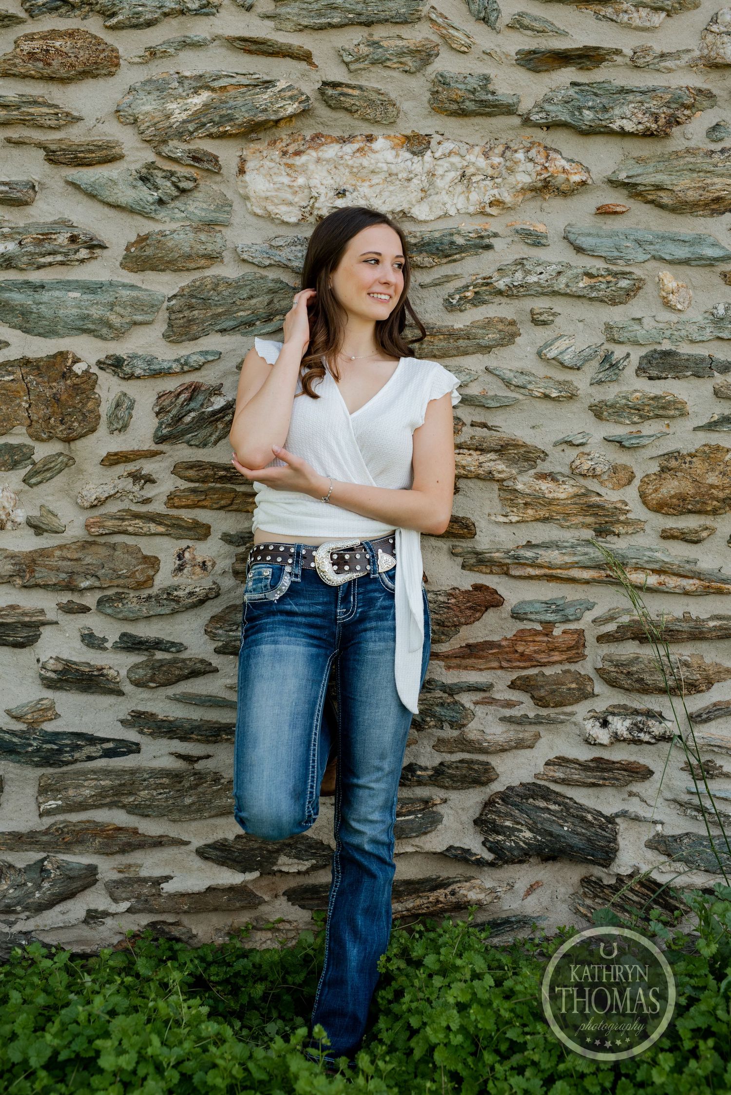 A woman in jeans and white top poses against a textured stone wall outdoors.