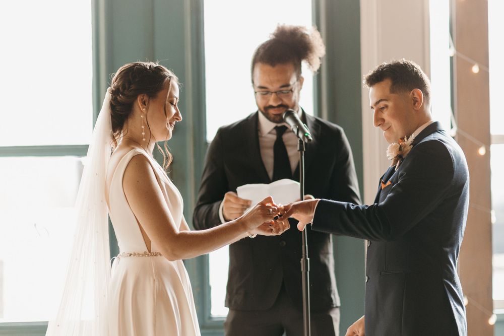 Bride places wedding ring on grooms finger during intimate indoor ceremony with officiant.