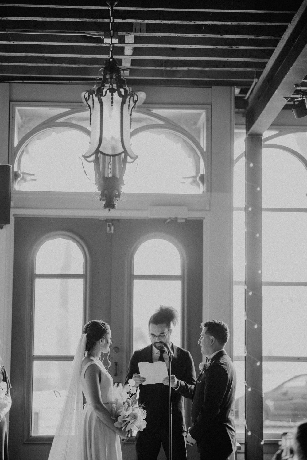 Black and white wide shot of wedding ceremony beneath ornate chandelier and arched windows.