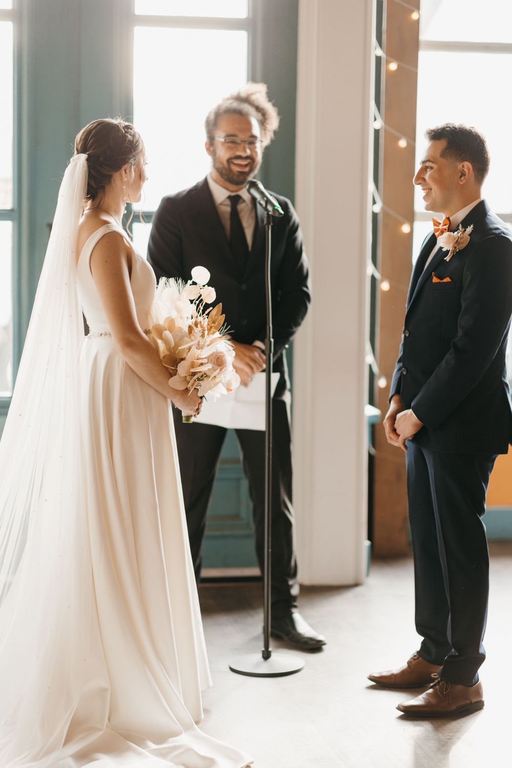 Bride and groom exchange vows at indoor wedding ceremony with officiant at microphone stand.