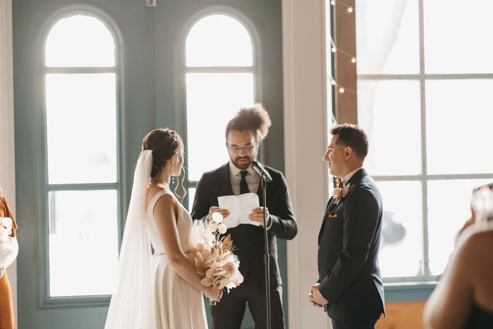 Smiling officiant reads ceremony script as bride holds bouquet facing groom indoors.