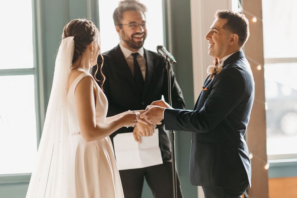 Bride and groom hold hands and smile during indoor wedding ring exchange ceremony.