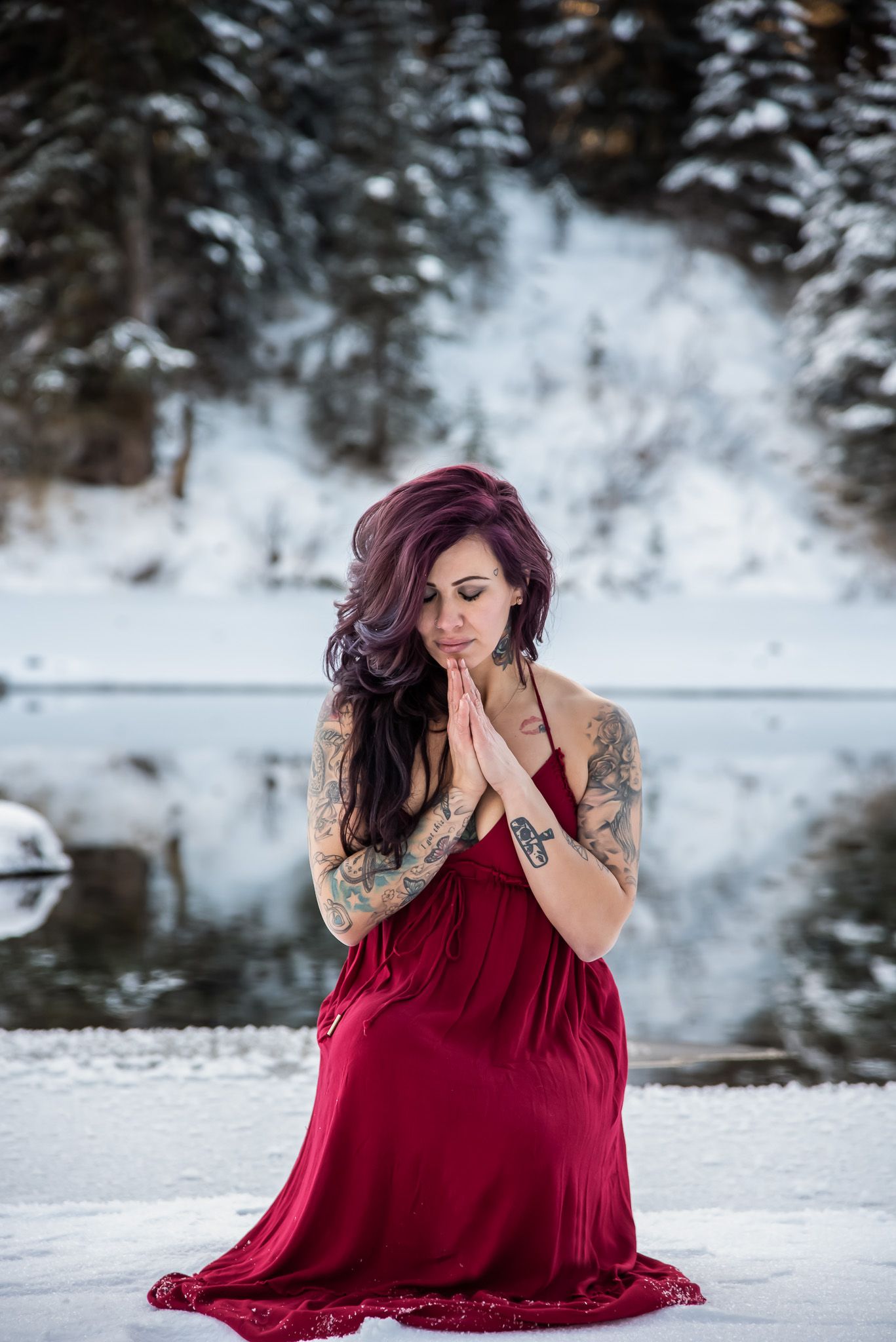 A person in a flowing red dress kneels in snow by a wintry lake with tattoos and burgundy hair.