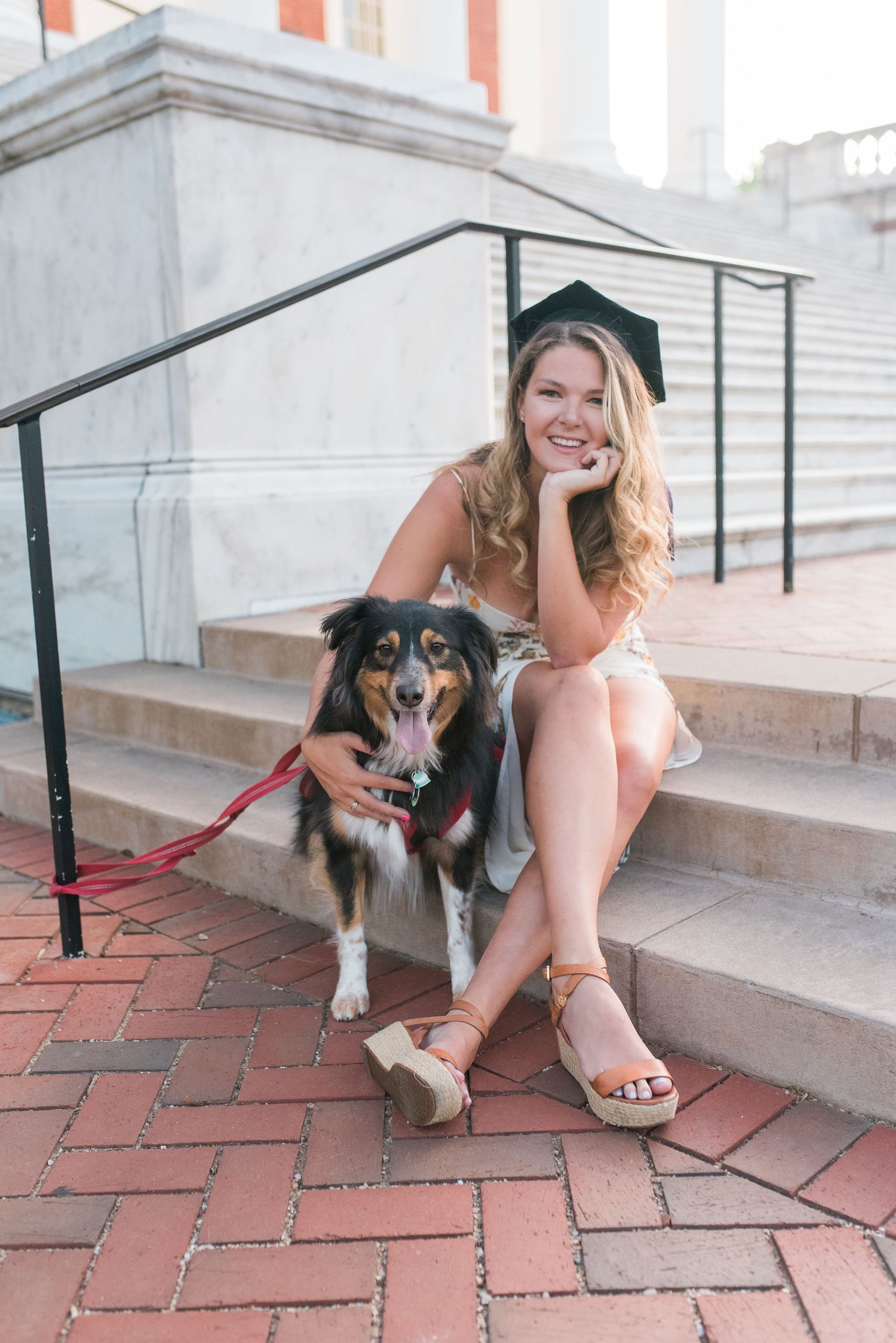 Graduate in a cap and gown sitting on brick steps with a happy Australian Shepherd dog.