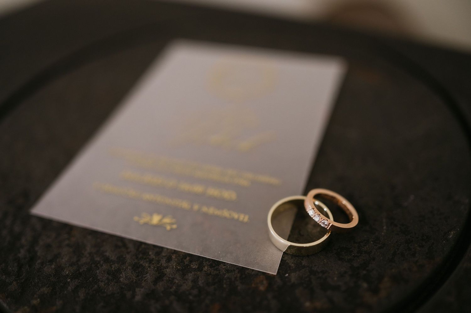 Two gold wedding rings sit on a dark invitation with gold lettering.