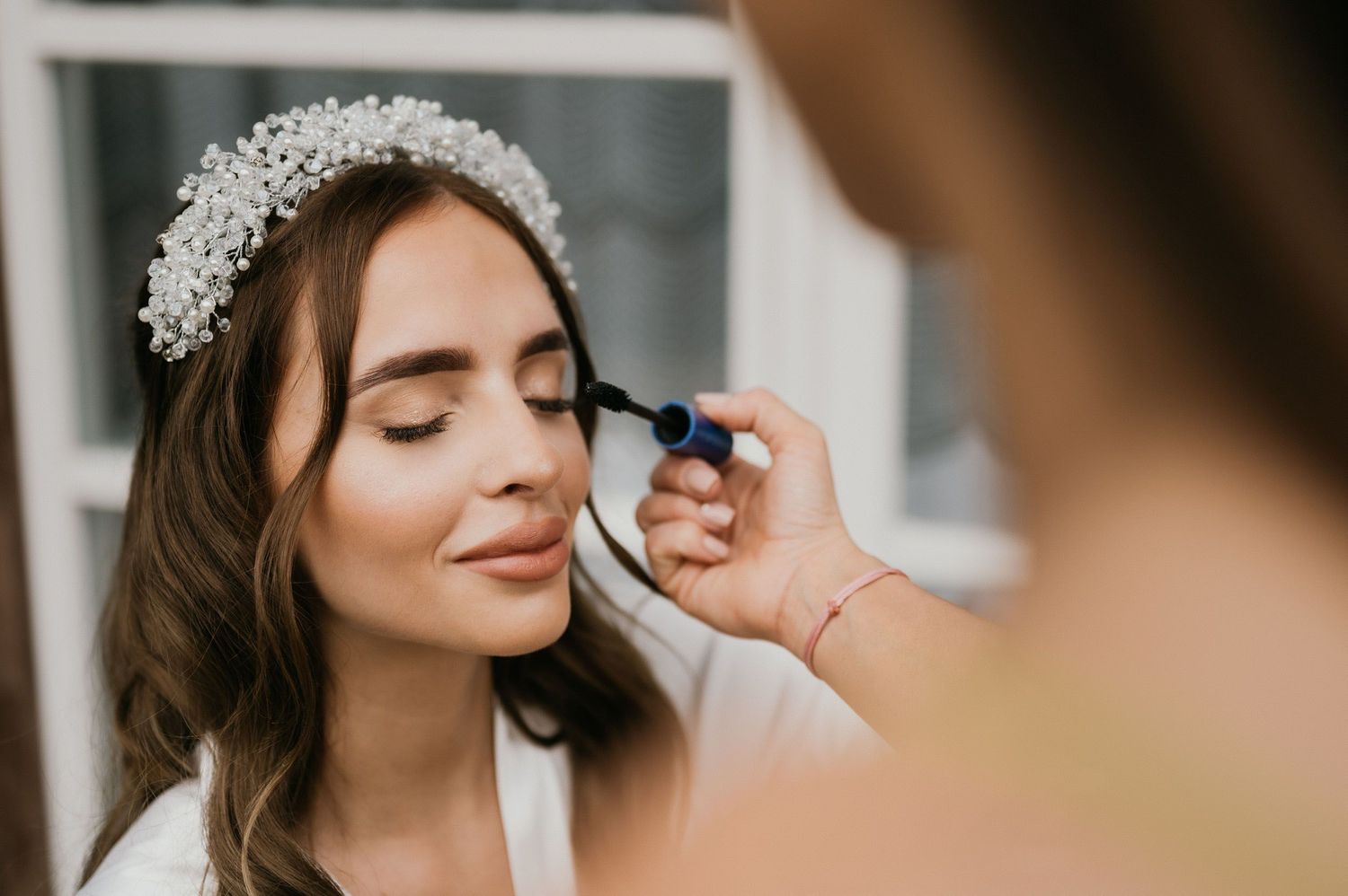 Makeup artist applies eyeshadow to bride wearing delicate crystal headpiece and white robe during wedding preparations.