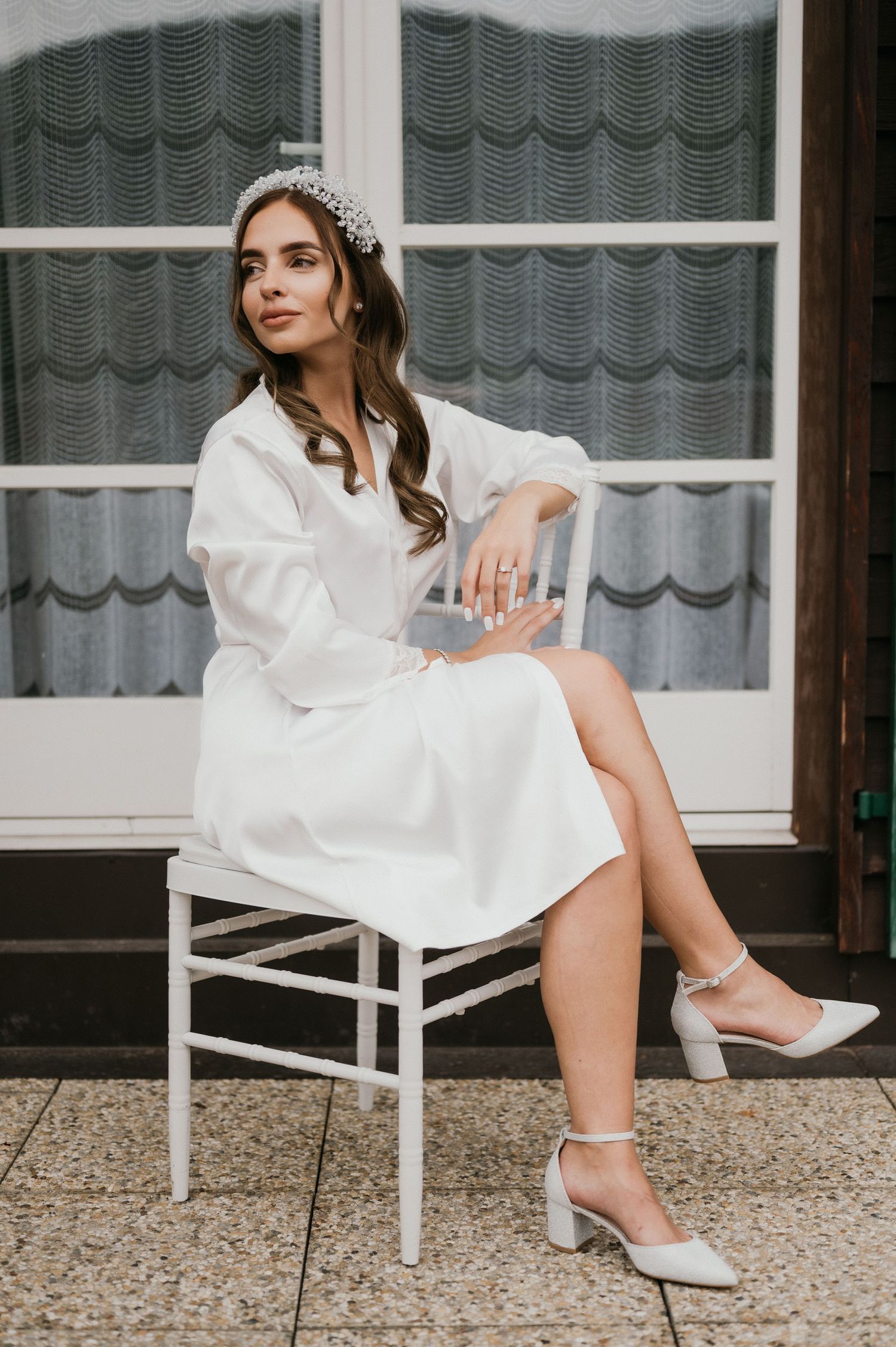 Elegant bride in white robe and crystal crown poses on white chair by window during getting ready session.