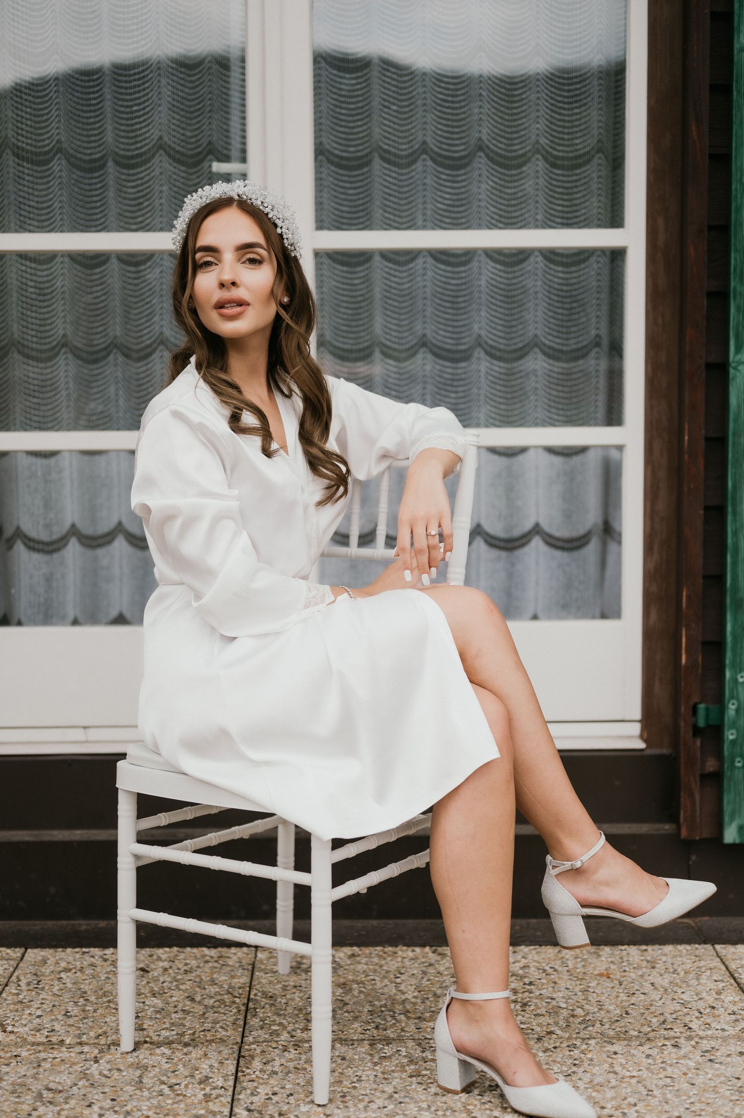 Bride in white silk robe and sparkly headpiece sits gracefully on chair during pre-wedding photoshoot.
