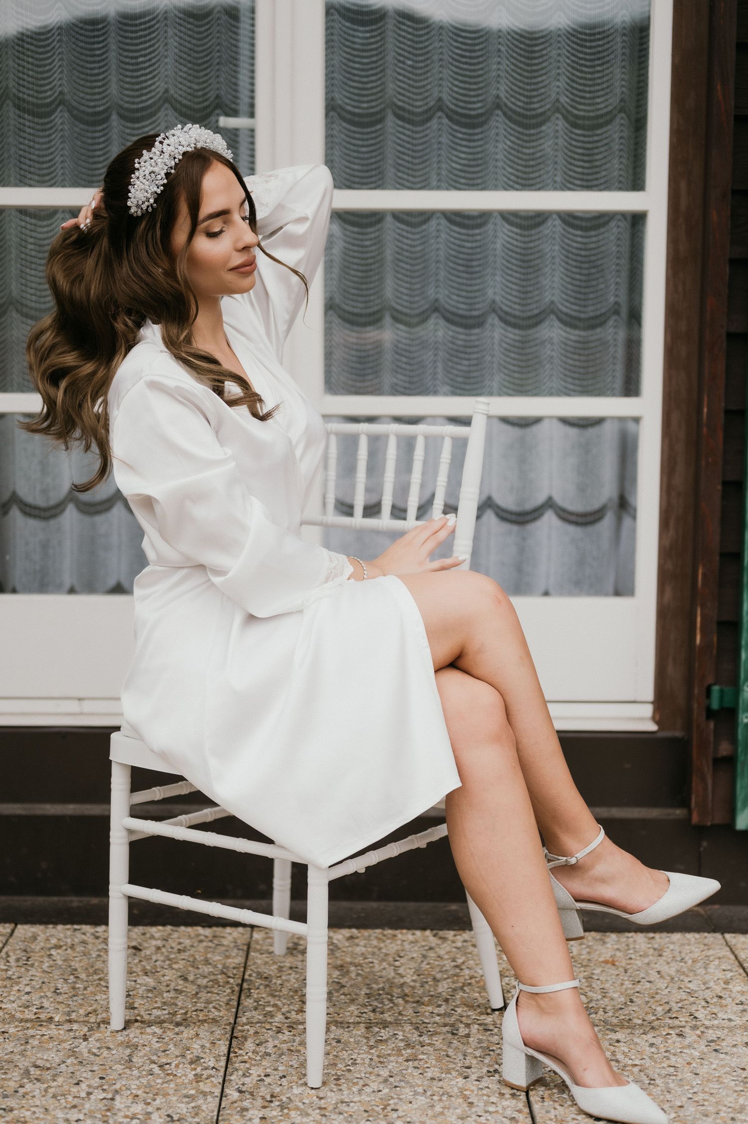 Candid bridal portrait showing white robe, crystal headpiece and silver heels against window backdrop.
