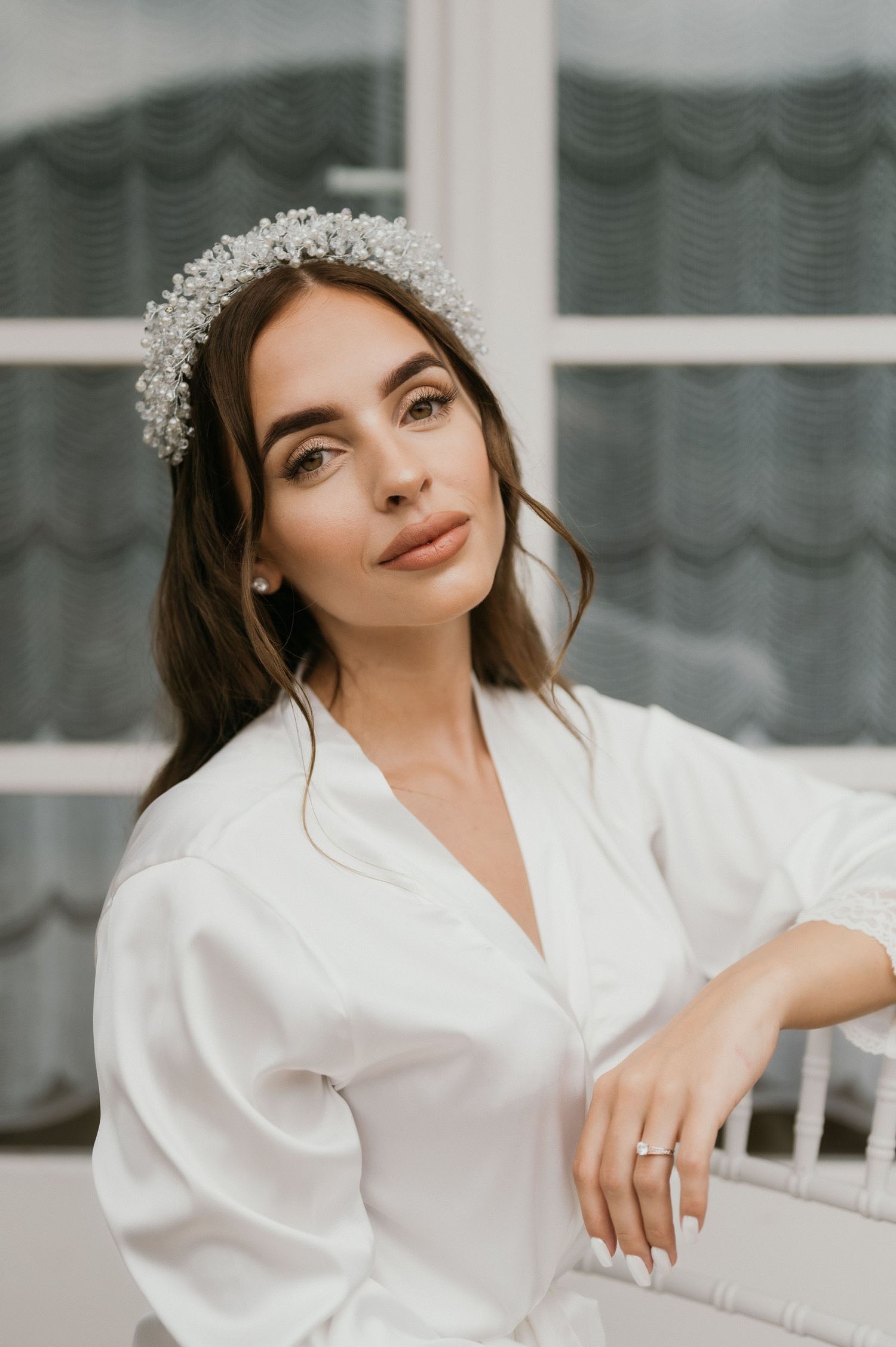Elegant bridal portrait featuring crystal crown and white silk robe against window background.
